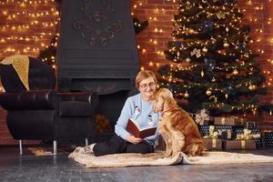 abuela con libro en la mano adentro con perro en la habitación decorada con navidad foto