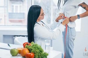 Female nutritionist measuring waist of patient by tape indoors in the office photo