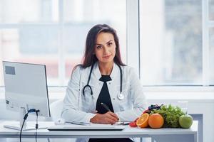 Female nutritionist in white coat sitting indoors in the office at workplace photo
