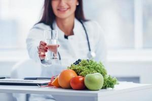 nutricionista femenina con bata blanca sentada en el interior de la oficina en el lugar de trabajo con un vaso de bebida foto