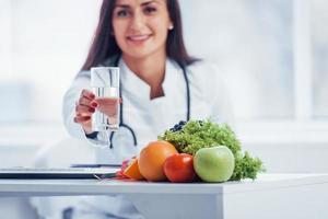 Female nutritionist in white coat sitting indoors in the office at workplace with glass of drink photo