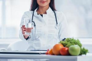 Female nutritionist in white coat sitting indoors in the office at workplace with glass of drink photo