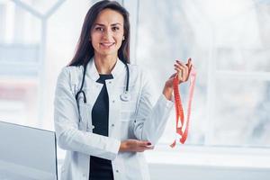 Female nutritionist in white coat standing indoors in the office at workplace with measuring tape in hands photo