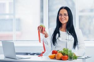 Female nutritionist in white coat holding apple with measuring tape photo