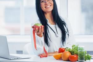 Female nutritionist in white coat holding apple with measuring tape photo