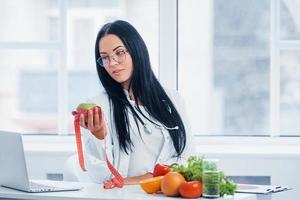 Female nutritionist in white coat holding apple with measuring tape photo