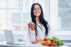 Female nutritionist in white coat holding apple with measuring tape photo