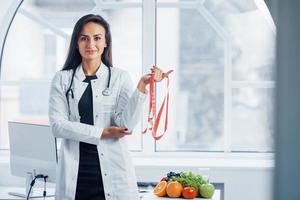 Female nutritionist in white coat standing indoors in the office at workplace with measuring tape in hands photo