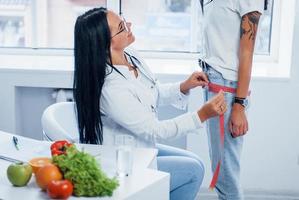 Female nutritionist measuring waist of patient by tape indoors in the office photo