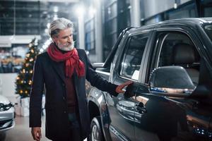 Fashionable old man with grey hair and mustache looking for a new car indoors in salon photo