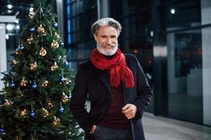 retrato de un anciano de moda con cabello gris y bigote parado cerca de un árbol decorado de navidad foto