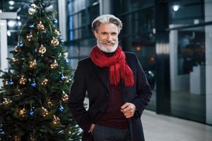 retrato de un anciano de moda con pelo gris y bigote parado cerca de un árbol decorado de navidad foto