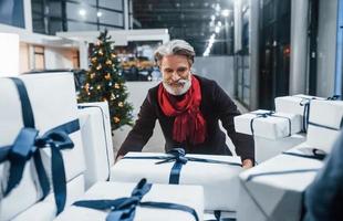 elegante anciano con cabello gris y bigote empacando regalos en cajas de regalo en el auto. árbol de navidad en el fondo foto