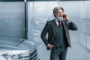 Senior businessman in suit and tie with gray hair and beard standing indoors with phone near car photo