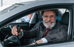 Modern stylish senior man with grey hair and mustache holding keys in modern car photo