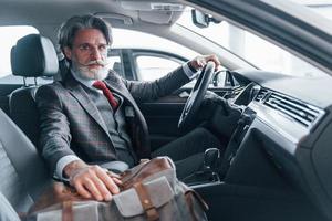 Fashionable senior man with grey hair and mustache on vacation with bag inside modern automobile photo