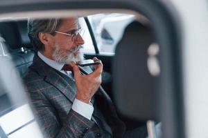 Modern stylish senior man with grey hair and mustache talking by the phone inside automobile photo