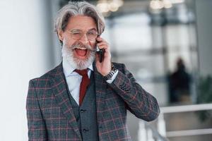 Senior businessman in suit and tie with gray hair and beard standing indoors with phone photo