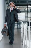 Senior businessman in suit and tie with gray hair and beard walking indoors with bag photo