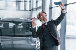 un anciano positivo con el pelo gris tomando selfie contra un coche moderno en el interior foto