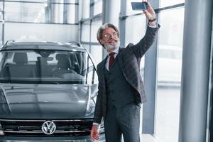 un anciano positivo con el pelo gris tomando selfie contra un coche moderno en el interior foto