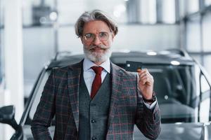 hombre con cabello gris y bigote parado contra un auto moderno en el interior con tarjeta de crédito en la mano foto