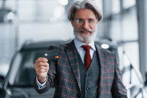 hombre con cabello gris y bigote parado contra un auto moderno en el interior con tarjeta de crédito en la mano foto