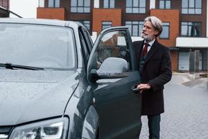 Fashionable senior man with gray hair and beard is outdoors on the street opens door of his car photo