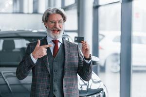 hombre con cabello gris y bigote parado contra un auto moderno en el interior con tarjeta de crédito en la mano foto