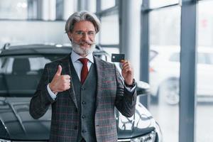 hombre con cabello gris y bigote parado contra un auto moderno en el interior con tarjeta de crédito en la mano foto