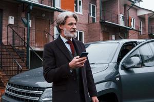 hombre mayor de moda con pelo gris y barba de pie con el teléfono cerca de su coche en la calle foto