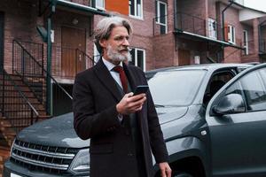 hombre mayor de moda con pelo gris y barba de pie con el teléfono cerca de su coche en la calle foto