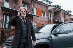 un anciano de moda con cabello gris y barba tiene una conversación por teléfono al aire libre en la calle cerca de su auto foto