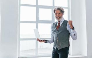 Hppy senior man in jacket and tie indoors in room against window with laptop in hands celebrating victory photo