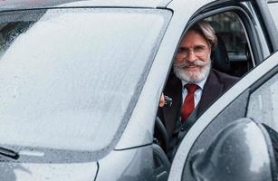 Happy stylish senior man with grey hair and mustache is in the modern car photo