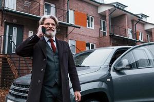 un anciano de moda con cabello gris y barba tiene una conversación por teléfono al aire libre en la calle cerca de su auto foto