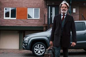 un anciano de moda con cabello gris y barba camina con una bolsa al aire libre en la calle cerca de su auto foto