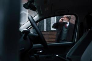 Fashionable senior man with gray hair and beard have conversation by phone outdoors on the street near his car photo