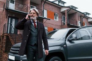 un anciano de moda con cabello gris y barba tiene una conversación por teléfono al aire libre en la calle cerca de su auto foto