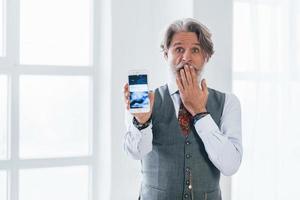 Shocked senior man in jacket and tie indoors in room against window showing display of smartphone photo