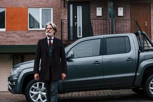 Fashionable senior man with gray hair and beard walks outdoors on the street near his car photo