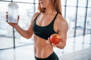 Sportive woman with nice body stands indoors with bottle of water and fresh apple in hands photo