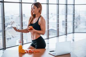 Sportive woman with nice body stands indoors with bottle of water and fresh apple in hands near table with laptop photo