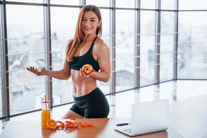 Sportive woman with nice body stands indoors with bottle of water and fresh apple in hands near table with laptop photo