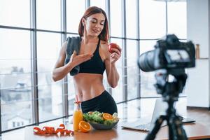 Female vlogger with sportive body standing indoors near table with healthy food photo