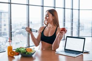 Sportive woman with nice body stands indoors with dumbbell and fresh apple in hands near table with laptop photo