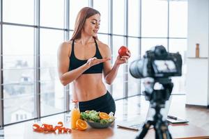 Female vlogger with sportive body standing indoors near table with healthy food photo