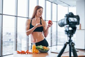 Female vlogger with sportive body standing indoors near table with healthy food photo