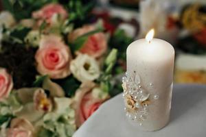 A beautiful white thick candle is burning, decorated with flowers and beads. Flowers in the background photo