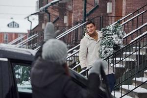 Man with christmas tree standing on the ladder of building and greeting female friend that is near car photo